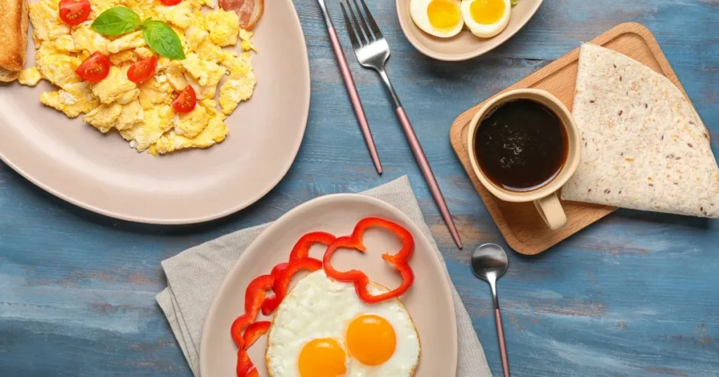 Breakfast spread featuring organic pasture raised eggs: scrambled eggs with cherry tomatoes and basil, sunny-side-up eggs with red bell pepper slices, boiled eggs, a cup of coffee, and a folded whole-grain tortilla on a blue wooden table.