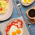 Breakfast spread featuring pasture-raised eggs: scrambled eggs with cherry tomatoes and basil, sunny-side-up eggs with red bell pepper slices, boiled eggs, a cup of coffee, and a folded whole-grain tortilla on a blue wooden table.