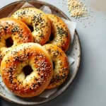 A plate of golden-brown gluten-free bagels topped with black and white sesame seeds, served with a side of creamy spread.