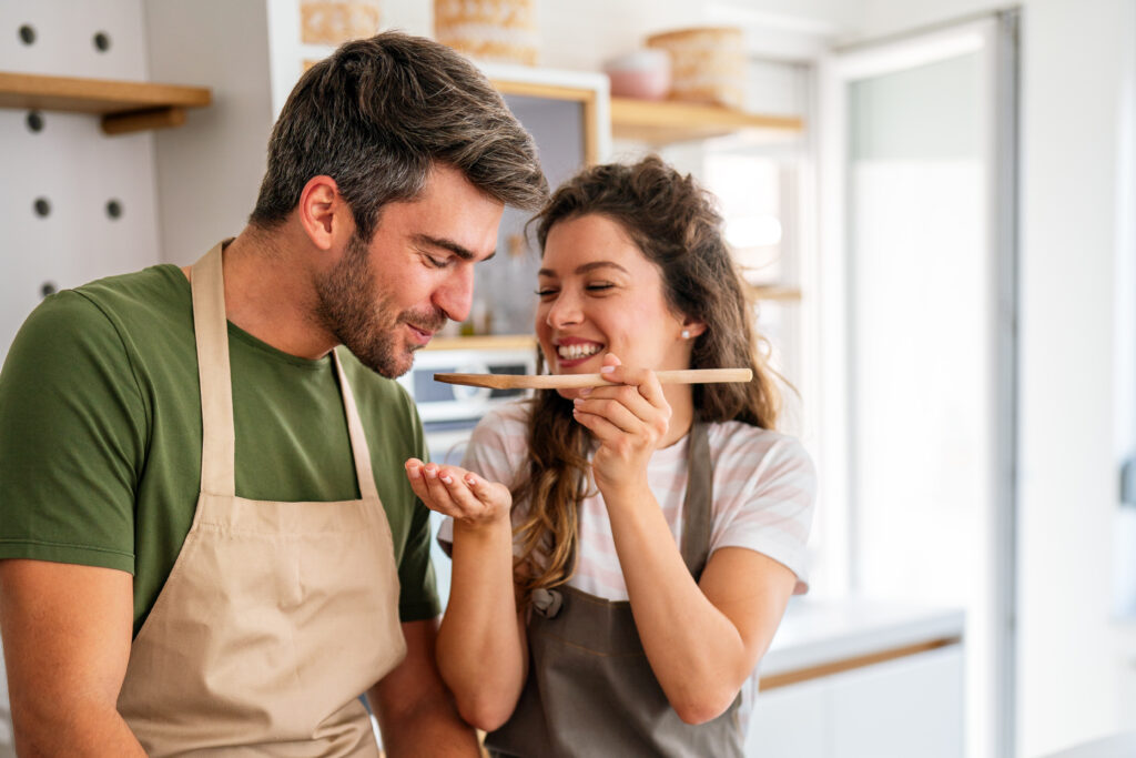 A couple wearing aprons cooks together in a bright, modern kitchen, tasting a homemade dish from a wooden spoon. The atmosphere is warm and inviting, perfectly capturing the spirit of “recipes yumz.”
