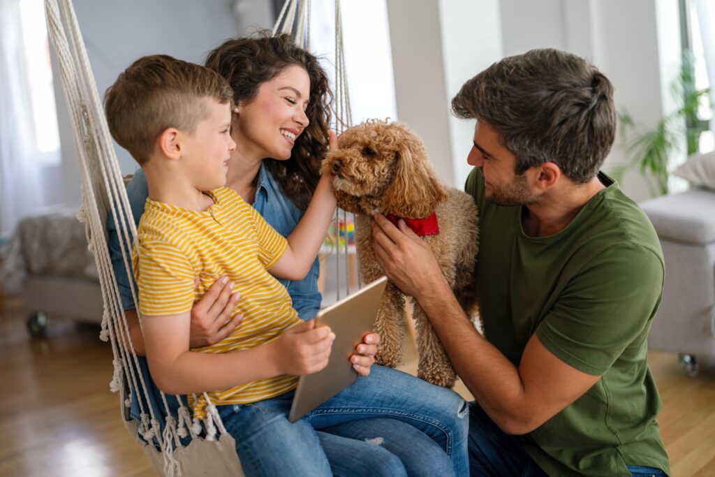 A family sits together on an indoor hanging chair in a bright living room. A young boy in a yellow striped shirt holds a tablet while an adult gently pets a small, curly-haired dog with a red collar. The mood is warm and joyful, perfect for sharing “recipes yumz” moments